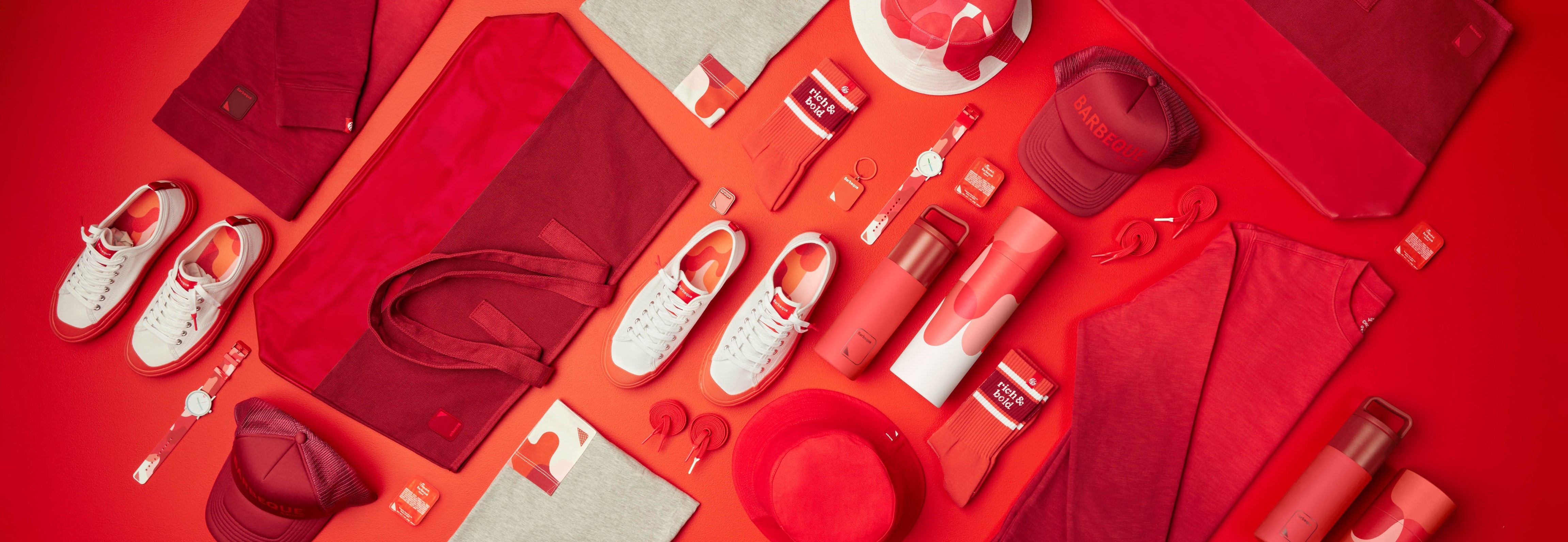 Overhead photograph of several red Barbeque Sauce Collection items, including water bottles, Dipped Kicks shoes, tote bags, pocket tees, crewnecks, and more