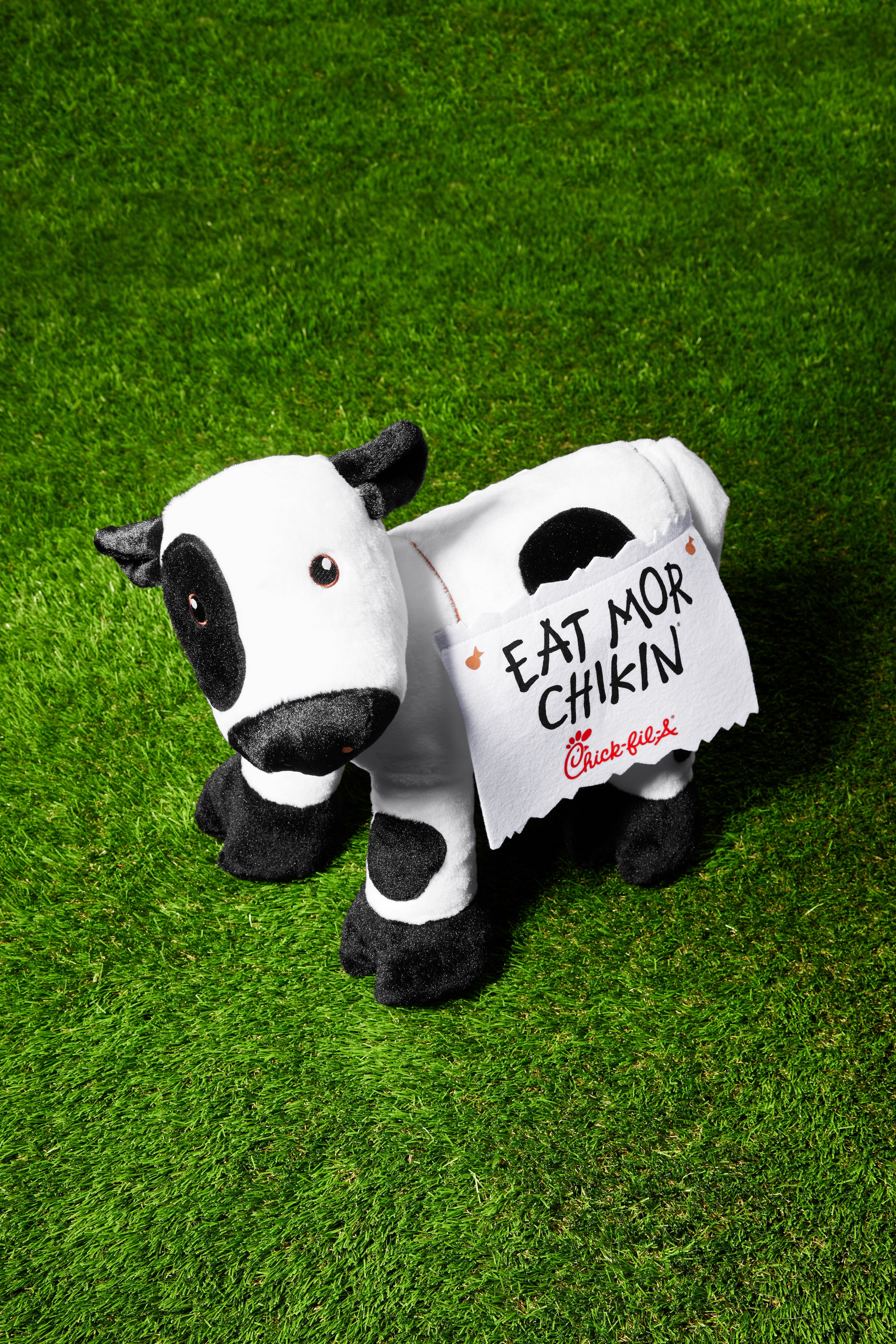 Black and white plush stuffed cow with decorative felt sign on his back that says “Eat Mor Chikin” on green turf