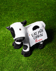 Black and white plush stuffed cow with decorative felt sign on his back that says “Eat Mor Chikin” on green turf