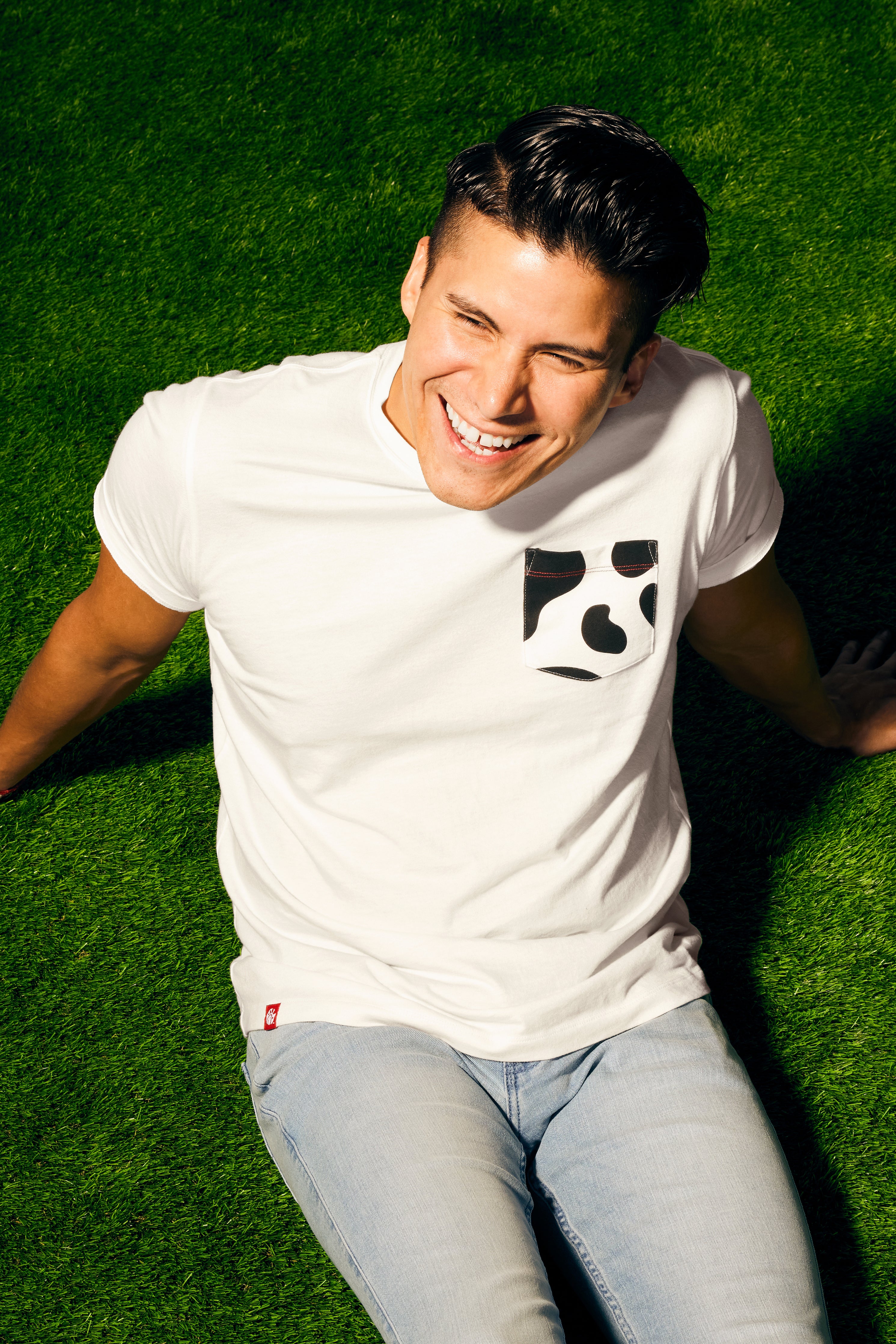 Man with brunette hair sitting on green turf wearing Cow Print Pocket Tee.