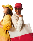 Woman wearing yellow Trucker Hat and yellow Tote Bag standing next to man with red Trucker Hat and red and canvas Tote Bag