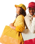 Woman wearing yellow Trucker Hat and yellow Tote Bag standing next to man with red Trucker Hat and red and canvas Tote Bag